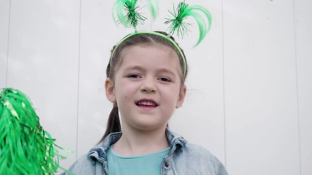 Beautiful happy kid dark hair and brown eyes with funny horns on the head looking at the camera, smiling and celebrating saint patrick's day waving with green pom poms white wall background