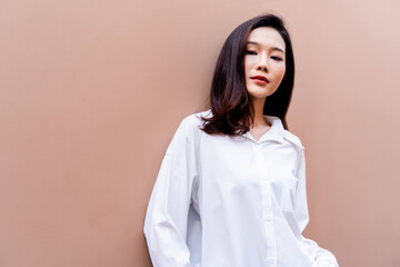 Portrait of a young asian girl with bright red lipstick and beautiful brown hair, and a gentle smile wearing a white shirt with open buttons, pressing her back against a desert sand colored wall.