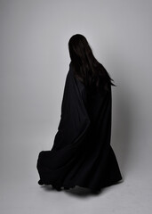 Full length portrait of pretty black haired woman wearing long dark gown nada cloak.  Standing pose facing away from the camera, against a  studio background.