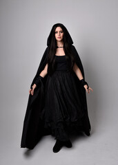 Naklejka premium Full length portrait of pretty black haired woman wearing long dark gown and a cloak. Standing pose facing away from the camera, against a studio background.
