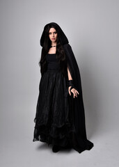 Naklejka premium Full length portrait of pretty black haired woman wearing long dark gown and a cloak. Standing pose facing away from the camera, against a studio background.