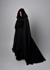 Naklejka premium Full length portrait of pretty black haired woman wearing long dark gown and a cloak. Standing pose facing away from the camera, against a studio background.
