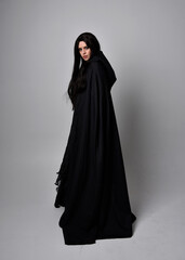 Full length portrait of pretty black haired woman wearing long dark gown and a cloak.  Standing...