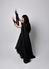 Naklejka premium Full length portrait of pretty black haired woman wearing long dark gown and a cloak. Standing pose facing away from the camera, against a studio background.