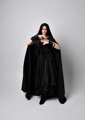 Full length portrait of pretty black haired woman wearing long dark gown and a cloak.  Standing pose facing away from the camera, against a  studio background.