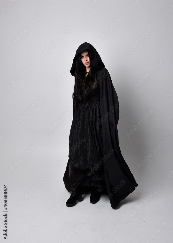 Poster Full length portrait of pretty black haired woman wearing long dark gown and a cloak.  Standing pose facing away from the camera, against a  studio background.