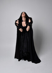 Naklejka premium Full length portrait of pretty black haired woman wearing long dark gown and a cloak. Standing pose facing away from the camera, against a studio background.