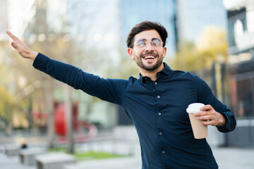 Young man holding a cup of coffee and calling a taxi outdoors.
