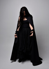 Full length portrait of pretty black haired woman wearing long dark gown and a cloak.  Standing...