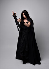 Full length portrait of pretty black haired woman wearing long dark gown and a cloak.  Standing pose facing away from the camera, against a  studio background.