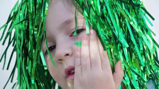 Adorable happy brown eyes child in green wig looking at the camera, hiding green star on her cheek, celebrating saint patrick's day, white wall background. Close up