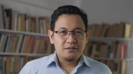 Young Asian man talking to camera while standing inside library, businessman doing video conference