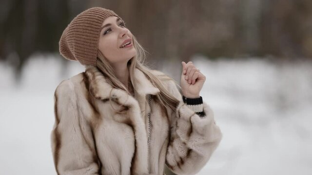 Portrait of an attractive fashionable blonde in a beautiful fur coat and a beige hat posing against the background of a winter park. The model is located on the street on a snowy background.