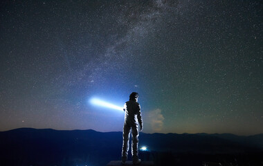 Cosmonaut wearing white space suit and helmet directs a blue ray of light into starry sky above...