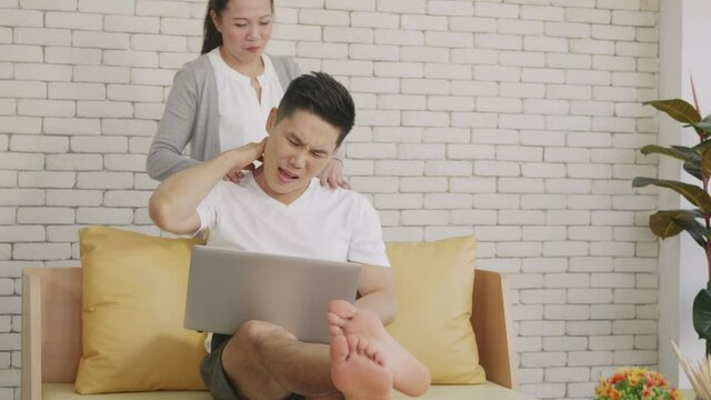 Happy Asian family couple husband and wife laughing sitting on sofa using laptop computer working from home Business man sitting on sofa home working on laptop her woman is giving her shoulder massag