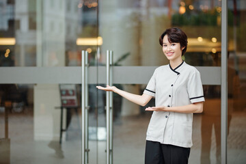 Happy young female concierge making welcoming gesture and inviting guests inside hotel - obrazy, fototapety, plakaty
