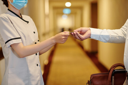 Close-up image of hotel manager in protective mask giving electronic key to man with big bag when they are standing in corridor