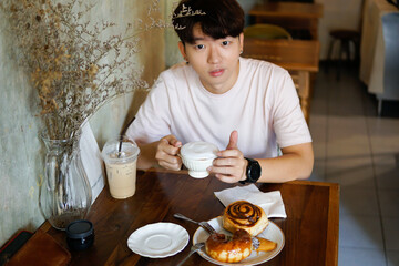 Handsome young asian man holding a cup of coffee and drinking coffee with bakery while sitting in the rest area of the coffee shop