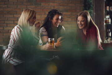 Three happy girlfriends are having fun time in pub. They are talking and drinking beer.