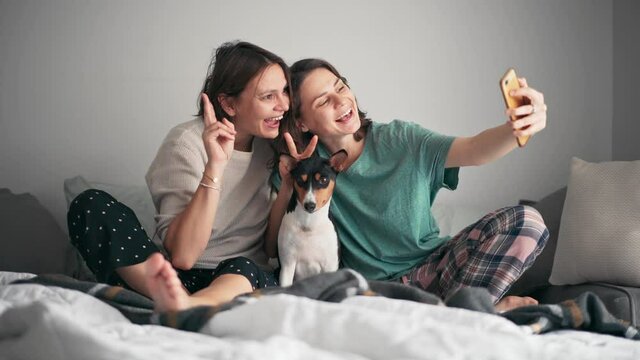 Beautiful lesbian couple taking a selfie with their cute basenji dog on a smartphone while sitting in pajamas on the bed. Cozy moments with a pet concept.