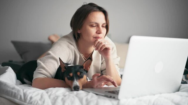 A middle-aged cheerful woman is working on her laptop while lying on the bed with her cute basenji dog.