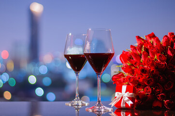 Two glasss of red wine and red roses bouquet with red gift box on table with colorful city bokeh lights for anniversary or Valentine's day concept.