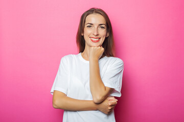 Fototapeta premium Young woman in white t-shirt holds hand under the chin, pink isolated background