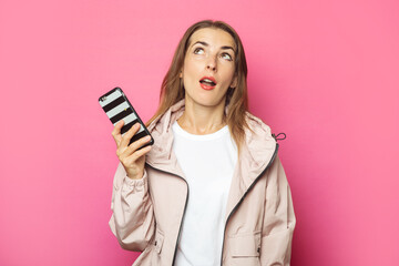 Young surprised woman holding a phone, pink isolated background