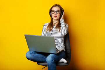 Young woman in headphones listening to music and sitting on chair and holding laptop on isolated yellow background