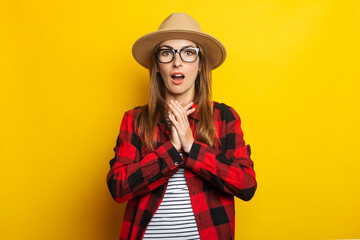 Young woman with surprised face in hat and plaid shirt on yellow background