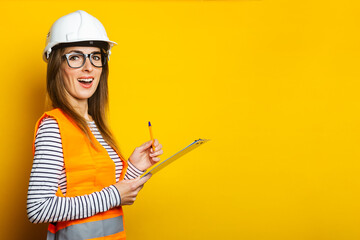 Fototapeta Young woman with a surprised face in a vest and hard hat holds a clipboard on a yellow background. Concept for construction, new building, renovation. Banner obraz