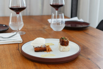 Gourmet dessert, including a honey cake, goat cheese and a honeycomb, served on a flat round stoneware plate with a tall rim. Background of a wood table, set with sharing plates, water and wine.
