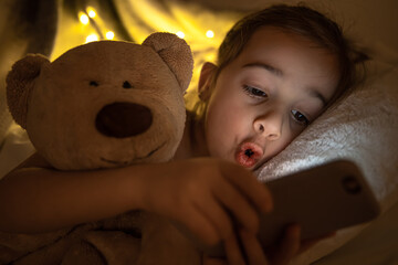 Little girl lies in bed and looks at the smartphone screen at night close up.