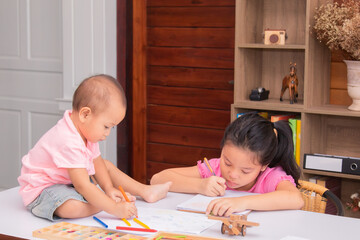 Adorable 2 kids sitting on desk full of crayon colors and white papers enjoy playing at home, 2 sibling share toys together, elder sister play with naughty younger sister after finished home work