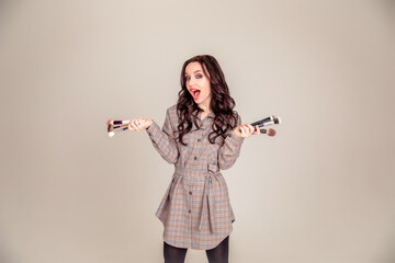 Young beautiful woman holding makeup brushes in her hands and posing on grey background at studio.