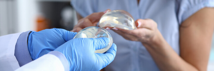 Patient of clinic hold breast implant in his hand and touch it with her finger. Doctor hold...