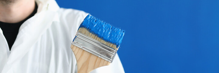 Man in white protective suit hold paintbrush in his hand. Wooden brush smeared with blue paint against blue wall.