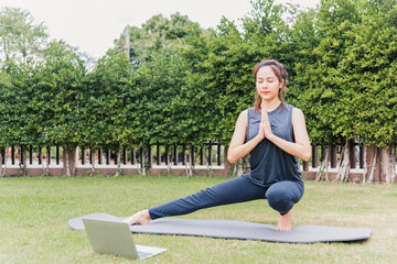 Fototapeta premium Asian young woman using laptop during online video training practicing yoga outdoors in meditate pose sitting on green grass in nature a field garden park, Stretching health care concept