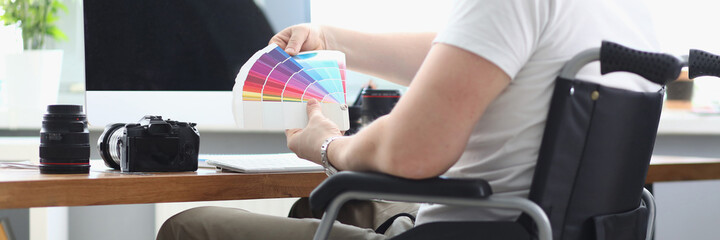 Man in white T-shirt and green pant sit in wheelchair and hold multi-colored palette in his hand. There is monitor, camera, len and keyboard on table.