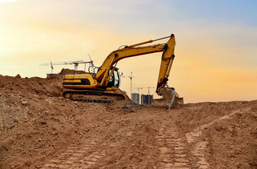 Fototapeta na wymiar Excavator digs dirt during roadwork at construction site. Heavy machinery and earth-moving equipment on road construction. Tower cranes in action on sunset background