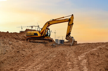 Excavator digs dirt during roadwork at construction site. Heavy machinery and earth-moving equipment on road construction. Tower cranes in action on sunset background