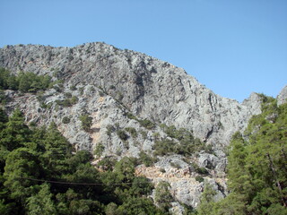 Panorama of steep insurmountable rocks surrounded by mountain forest at the foot of the clear blue sky.