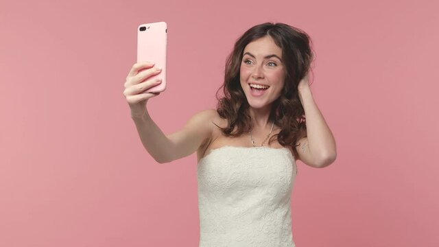 Funny bride young brunette woman 20s in beautiful white wedding dress posing isolated on pink background. Wedding concept. Doing selfie shot on mobile phone victory sign thumb up blowing send air kiss