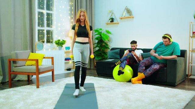 A woman doing lunges in a work out mat, there s two men behind her sitting on the couch and going on their phones, they re all in a modern room