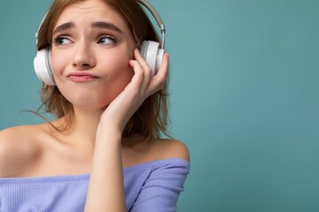 Closeup photo of attractive thoughtful young blonde woman wearing blue crop top isolated over blue background wall wearing white wireless bluetooth headphones listening to cool music and having fun