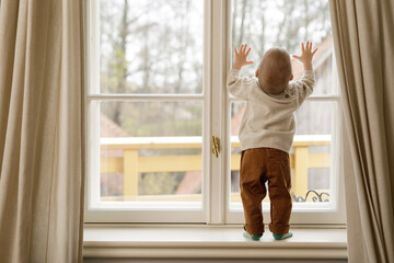 Baby boy is standing on the windowsill and looking at the window