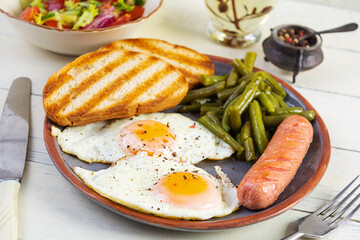 Breakfast with eggs, grilled sausage, green beans and toast on wooden background