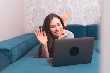 Photo of young woman smiling and talking with friends online on laptop while laying on sofa.