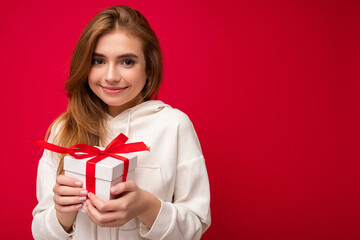 Beautiful happy young blonde woman isolated over colourful background wall wearing stylish casual clothes holding gift box and looking at camera