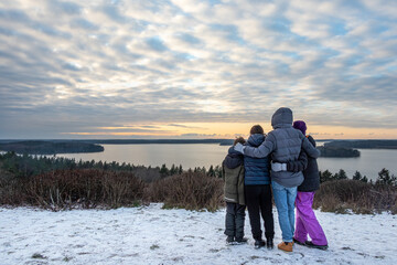 Family together holding each other and looking at a view. Mountain top winter sunset snow scene with water and horizon.